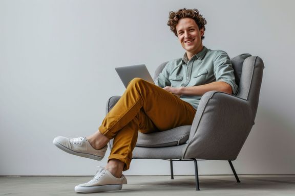 junger Mann sitzt mit einem Laptop in einem großen, gemütlich wirkenden, grauen Sofasessel