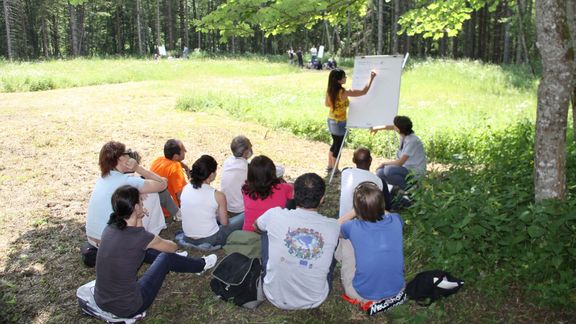 Naturschutzfachkraft Ausbildung im Freien: Studierende sitzen auf einer Wiese unter einem Baum und blicken zur Vortragenden, die gerade etwas auf ein Flipchart schreibt