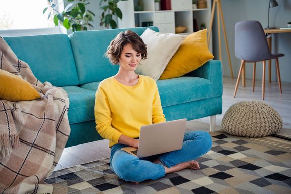 Junge Frau im gelben Pullover sitzt mit Laptop am Teppich und lehnt an der Couch
