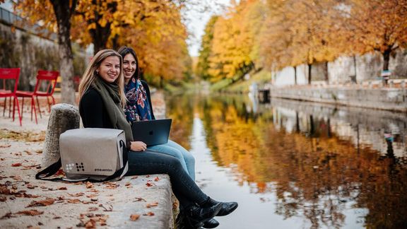 2 Studierende sitzen in herbstlicher Landschaft am Fluss
