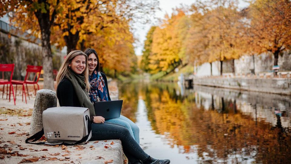 2 Studierende sitzen in herbstlicher Landschaft am Fluss 