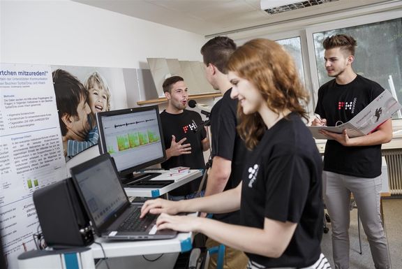 Forschung Fachhochschule Kärnten - CONNA zeigt eine Studierende vor dem Computer, während im Hintergrund drei weitere Studenten miteinander interagieren.