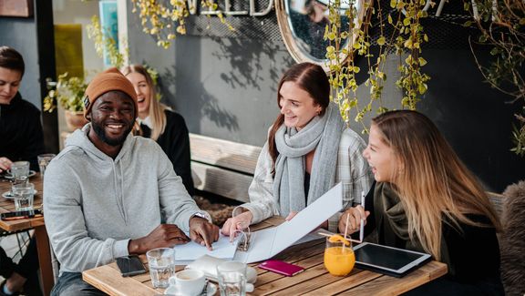 Drei Studierende sitzen in einem Cafe an einem Tisch im Freien mit Lernunterlagen